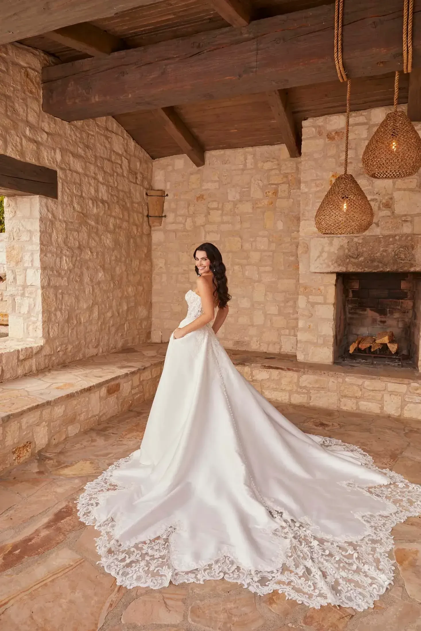 Bride in elegant satin gown with lace train, standing in rustic stone room with wooden beams and lit pendant lamps, evoking a warm, romantic ambiance.