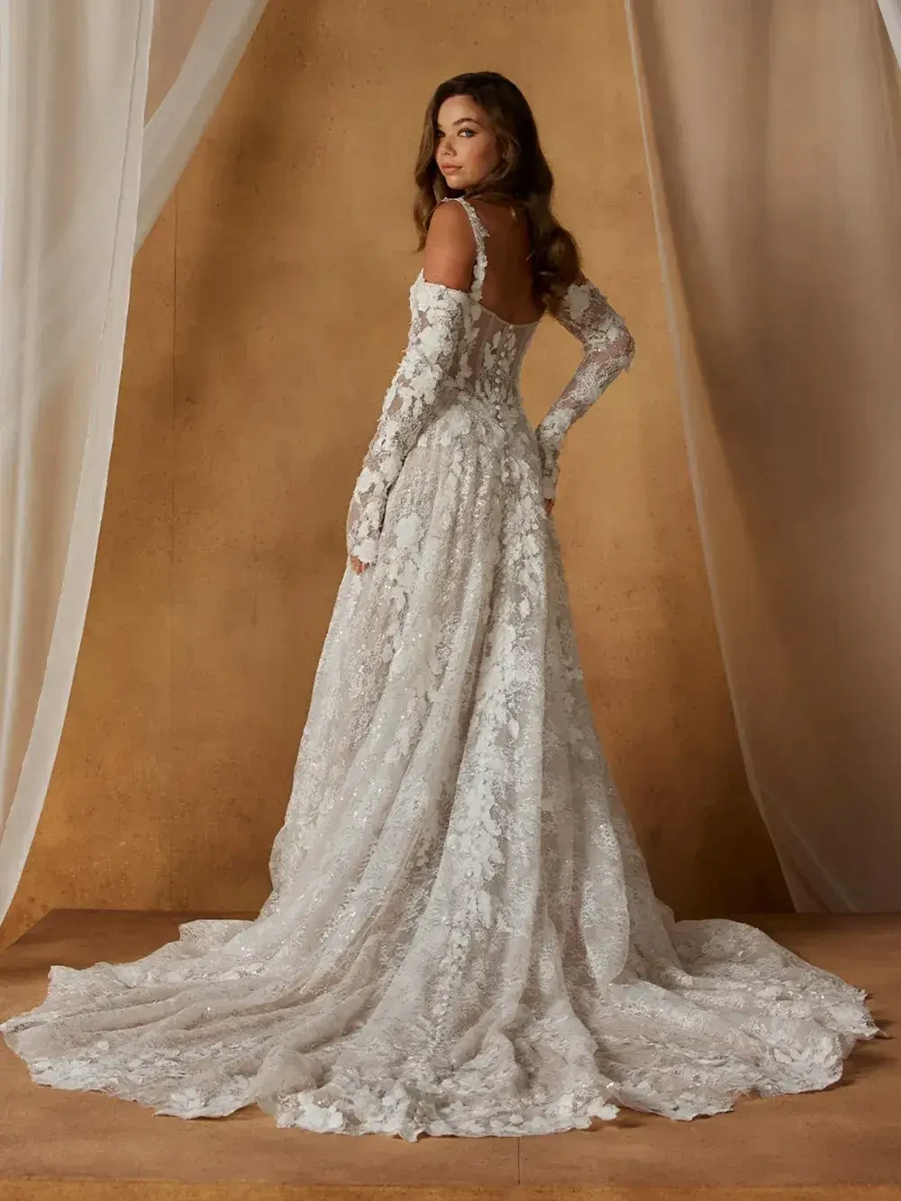 A woman in a long, lace wedding dress with a train and off-the-shoulder sleeves stands gracefully before beige drapes, exuding elegance and romance.