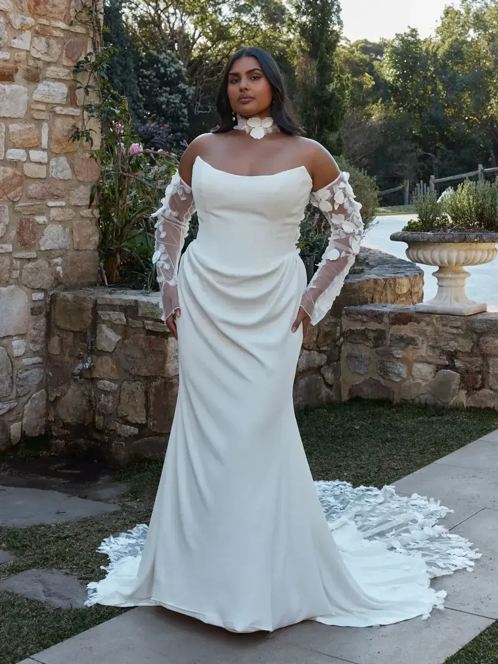 A woman in a white wedding dress with sheer, floral-patterned sleeves stands confidently in a garden. Stone walls and greenery create a serene backdrop.