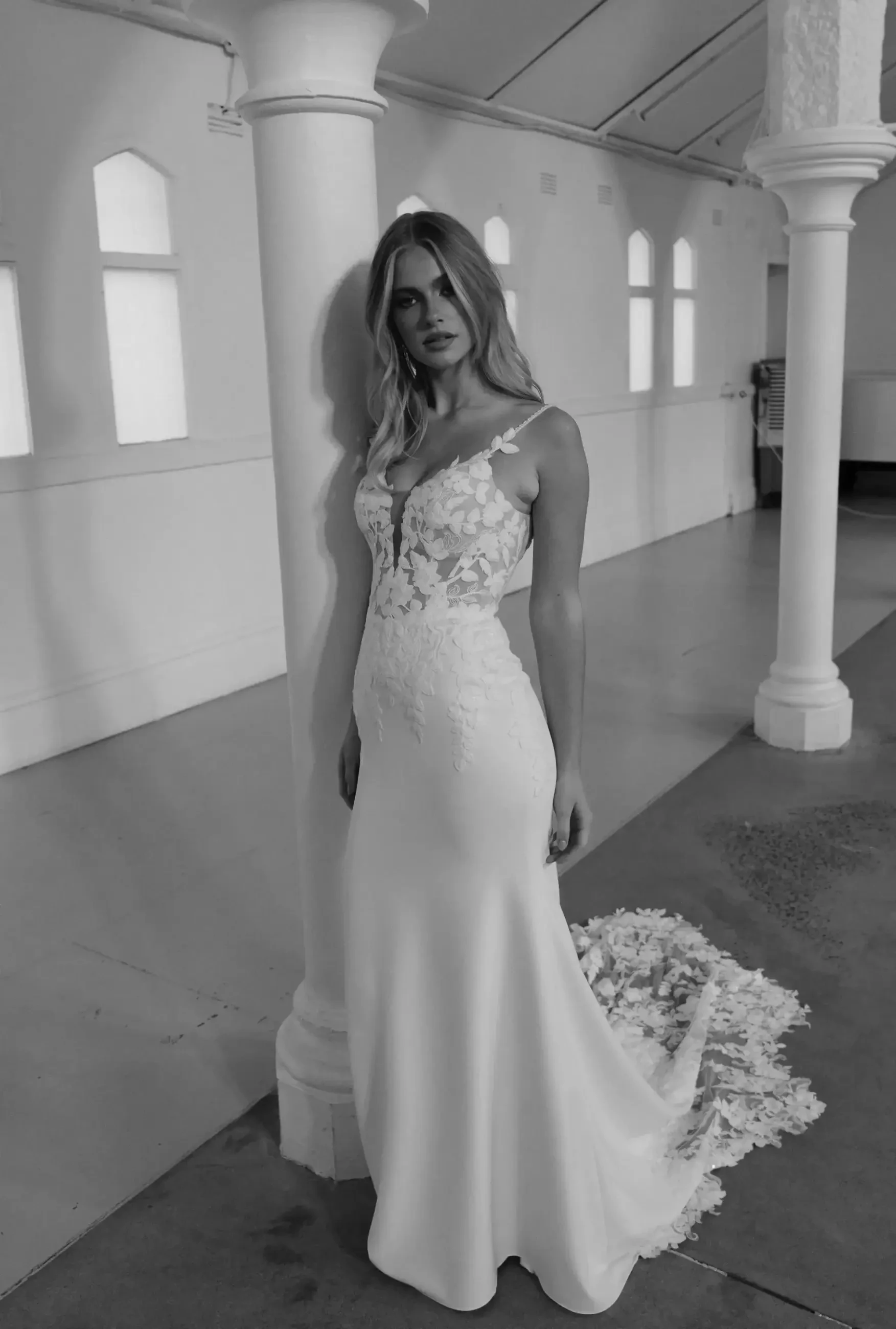 A woman in an elegant lace wedding gown with a long train stands by a pillar in a softly lit hall. Her expression is serene and poised.
