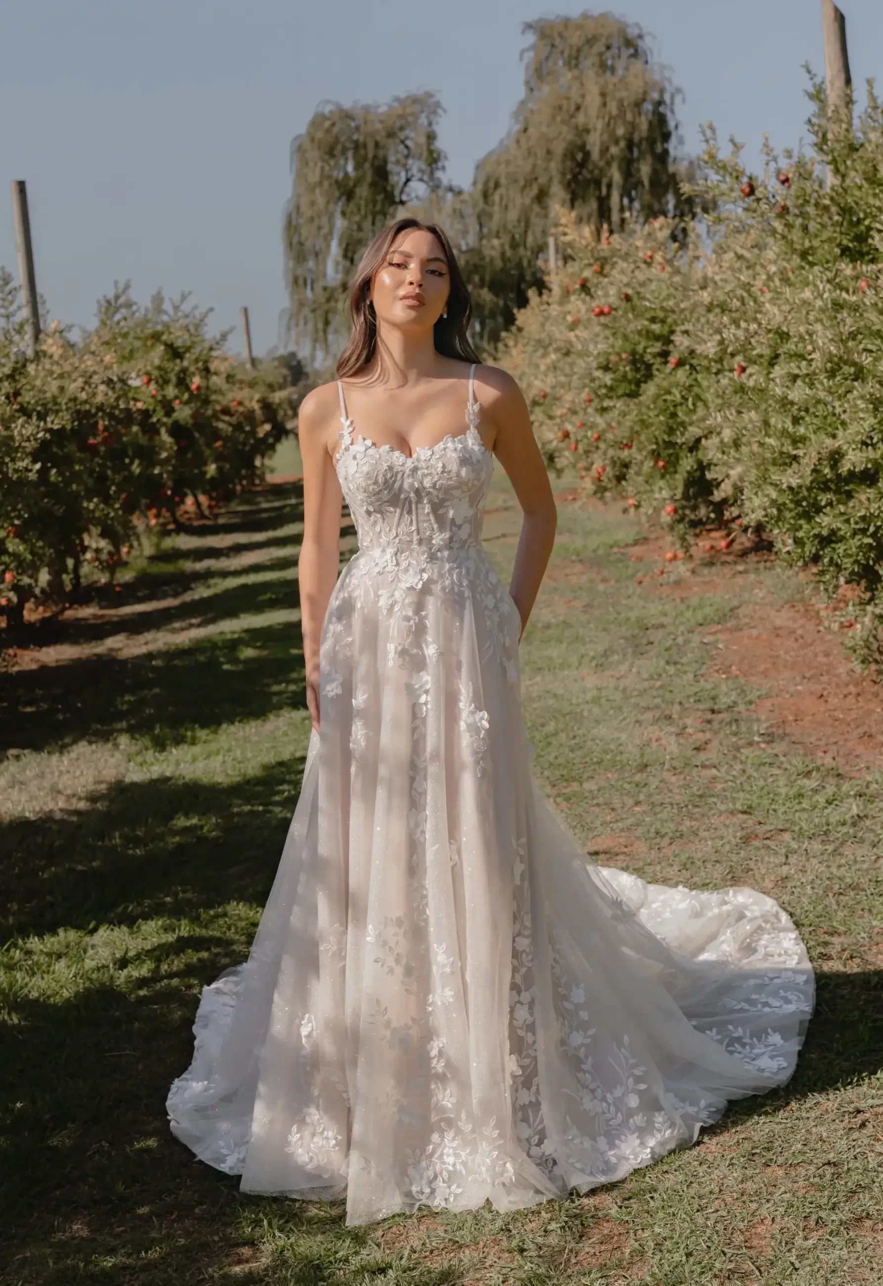 A woman in a lace wedding dress stands in a sunlit orchard. She has long hair and a serene expression. Green foliage and blue sky are in the background.