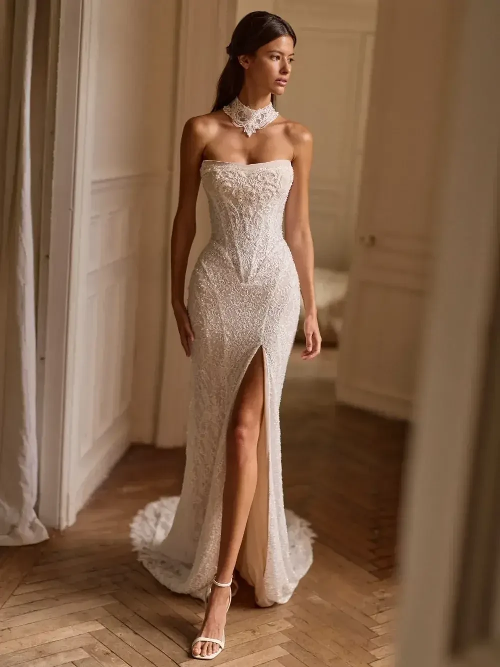 A model wearing a white strapless gown with intricate lace details and a high slit, walking in an elegantly decorated room.