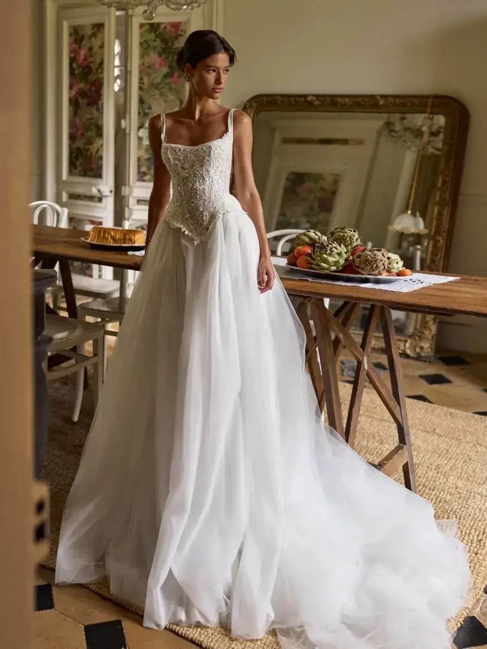 A woman in an elegant white lace and tulle wedding dress stands in a vintage room with floral wallpaper. The table holds fruit, suggesting a calm, romantic atmosphere.