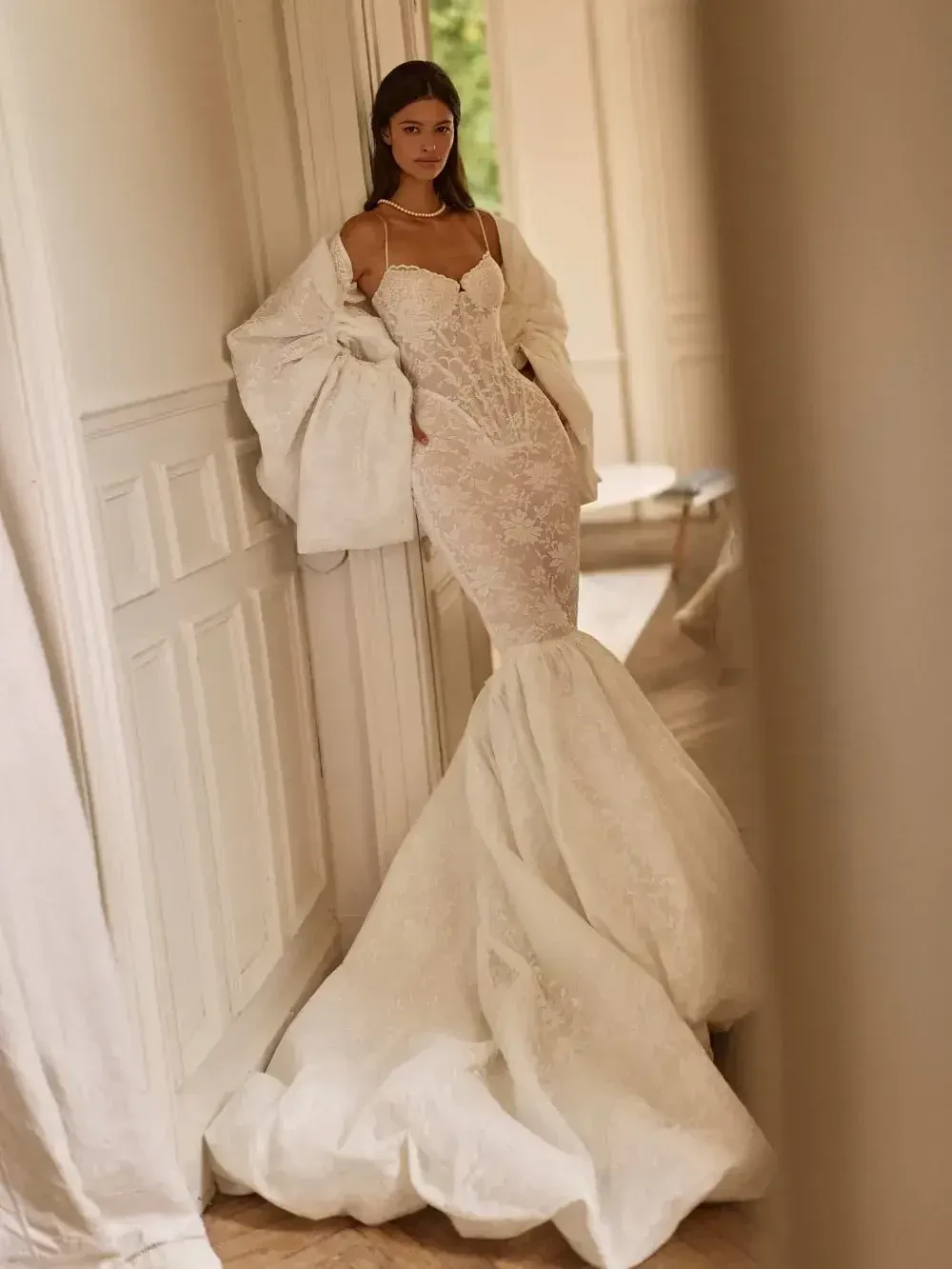 A woman wearing an elegant, form-fitting lace wedding dress with a mermaid silhouette and puffy train stands against a white paneled wall, exuding sophistication.