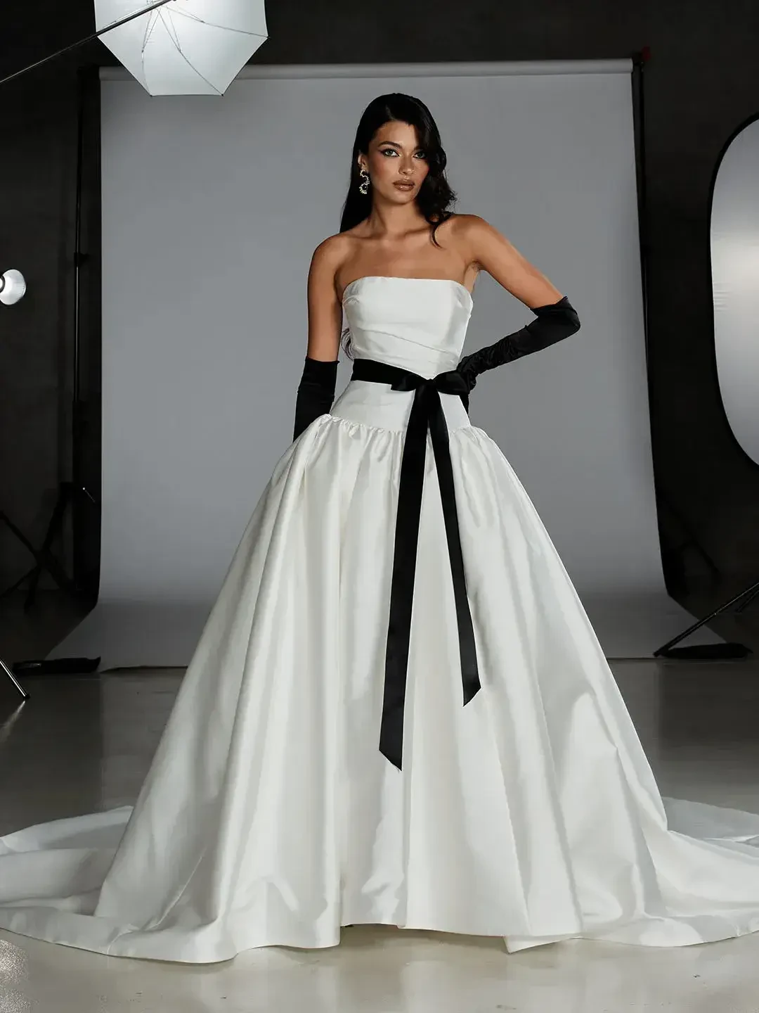 A model wearing a strapless white gown with a black sash and long black gloves, standing in a studio setting.