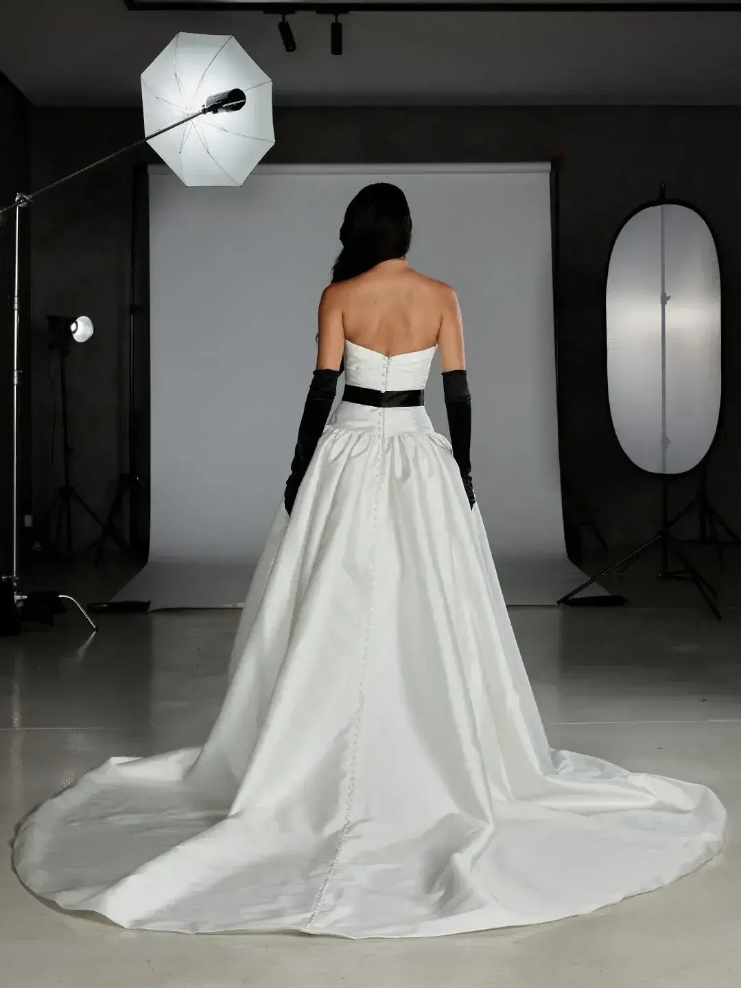 A woman in a white gown stands with her back to the camera, showcasing a long train, with black gloves on her arms and a studio lighting setup in the background.