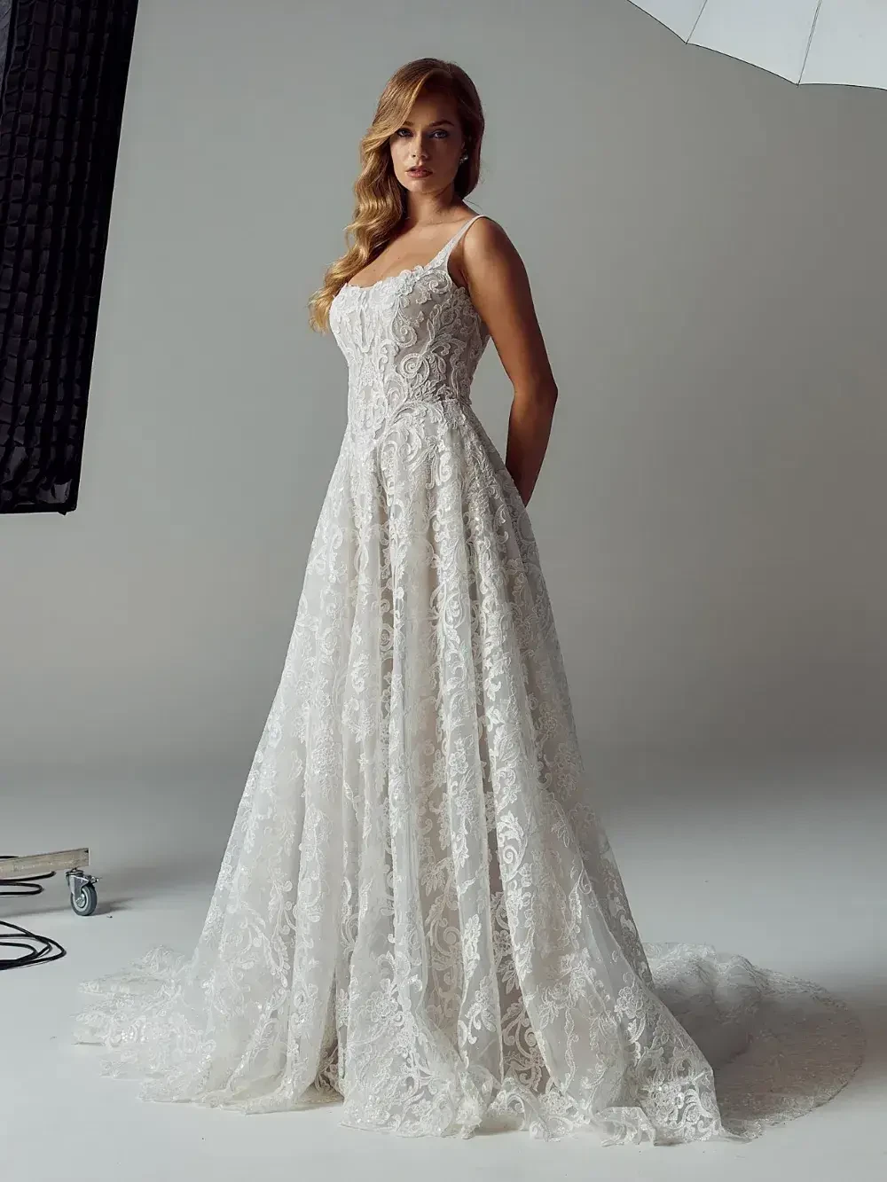 A woman in a lace wedding dress standing in a studio with neutral background lighting.