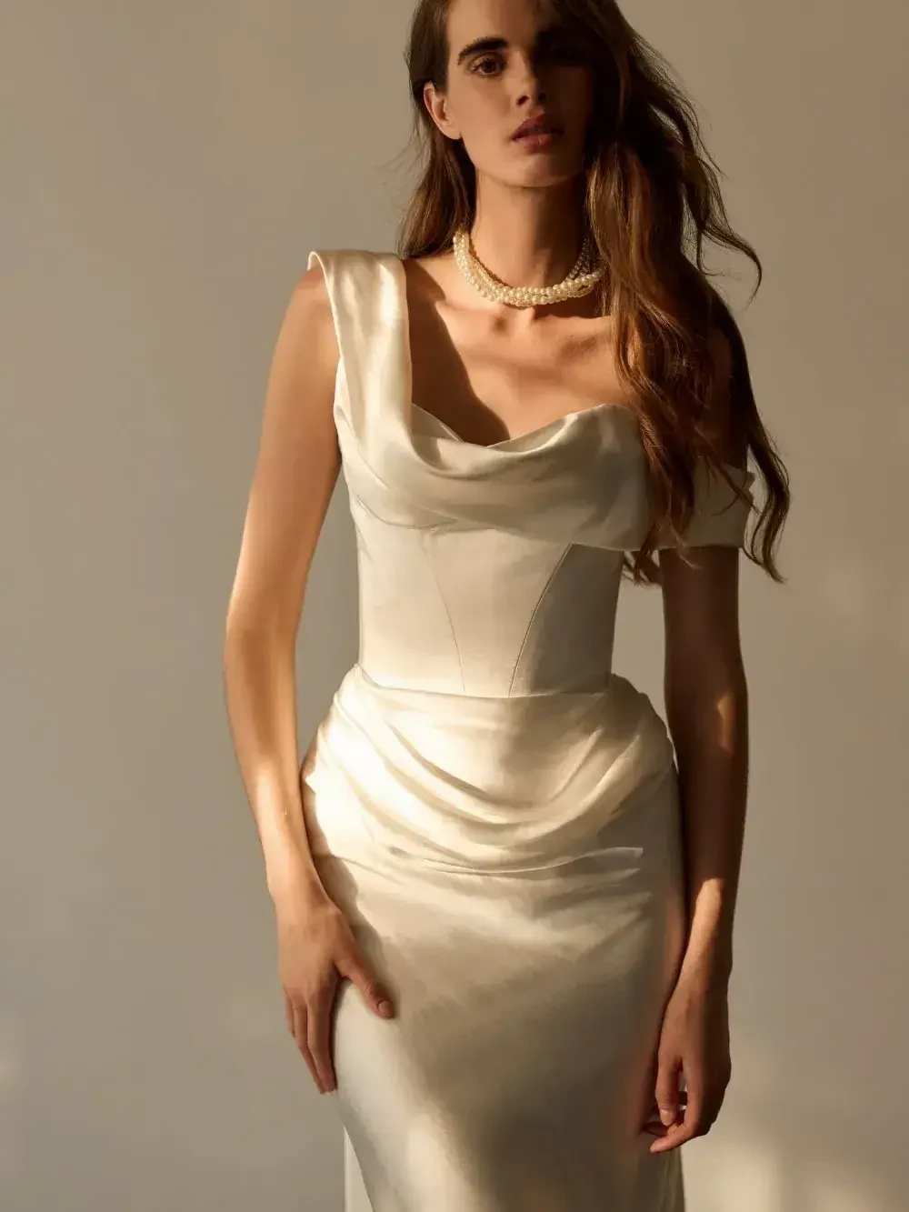 A woman in a fitted, off-white dress with short sleeves and a draped neckline, standing against a neutral background. She has long, flowing hair and is wearing a pearl necklace.