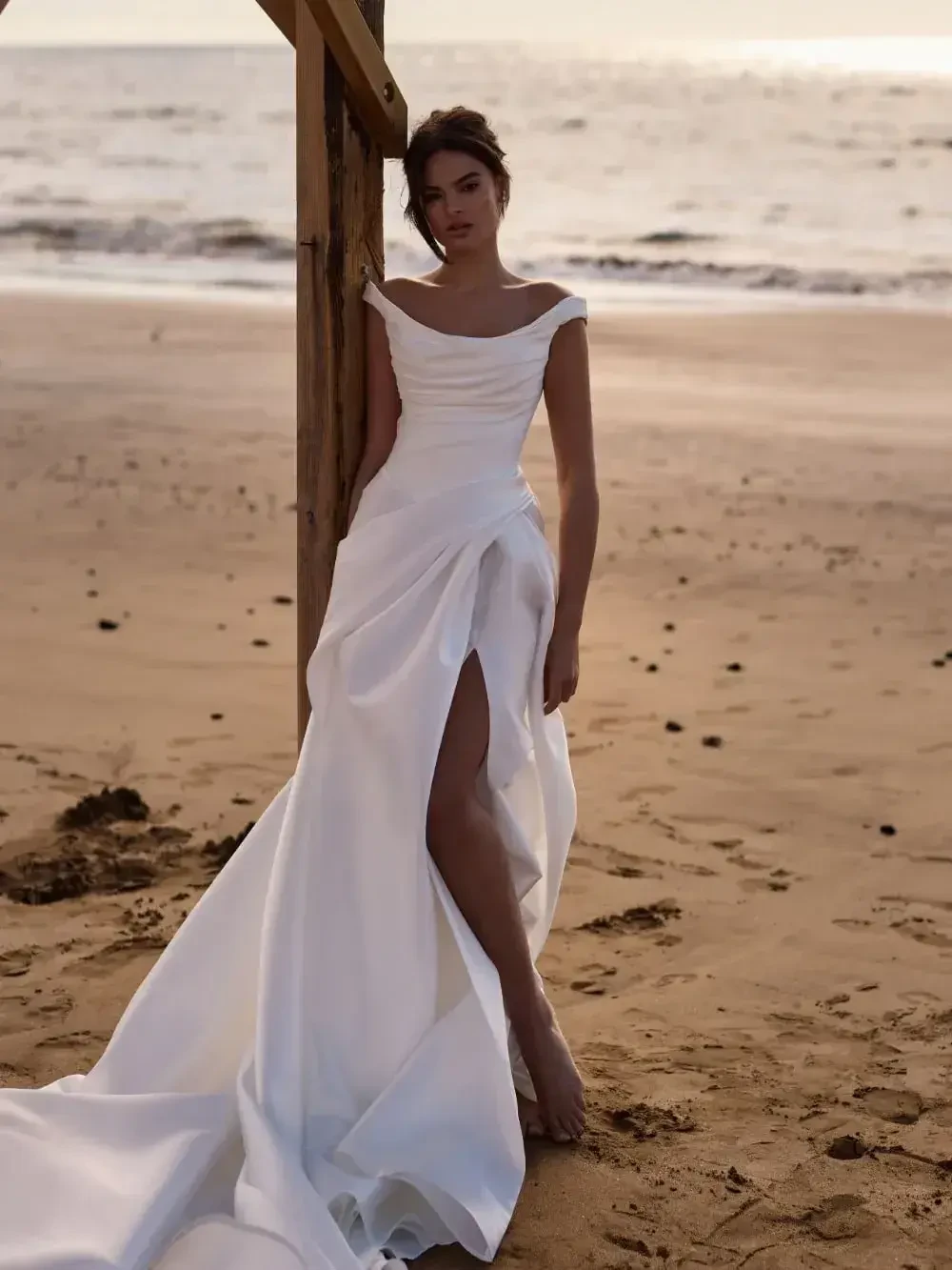 A woman wearing an elegant white dress with a high slit stands on a beach, leaning against a wooden structure with the ocean in the background.