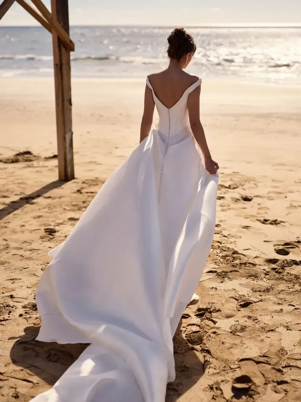 A woman in a white wedding dress walking away on a sandy beach with the ocean in the background.