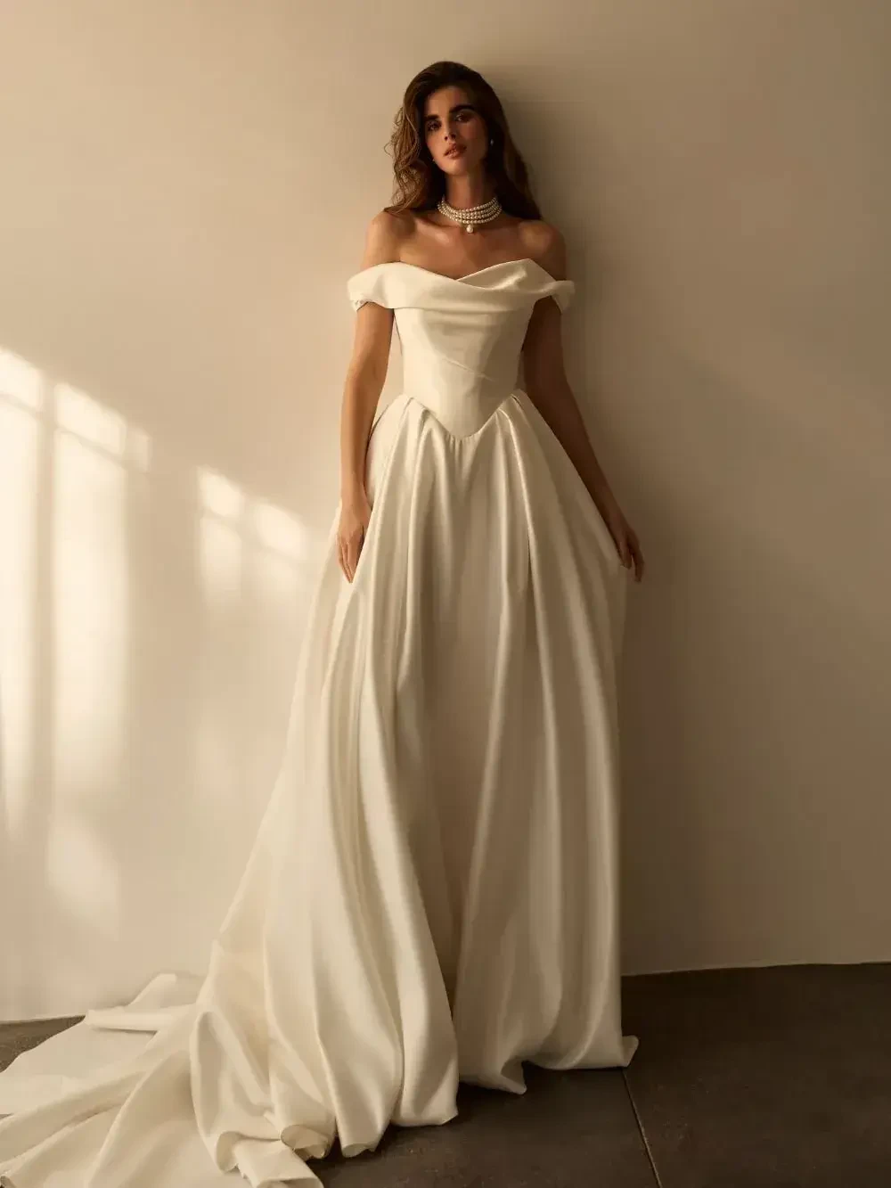 A model wearing an elegant off-the-shoulder white gown, standing against a plain wall with soft lighting.