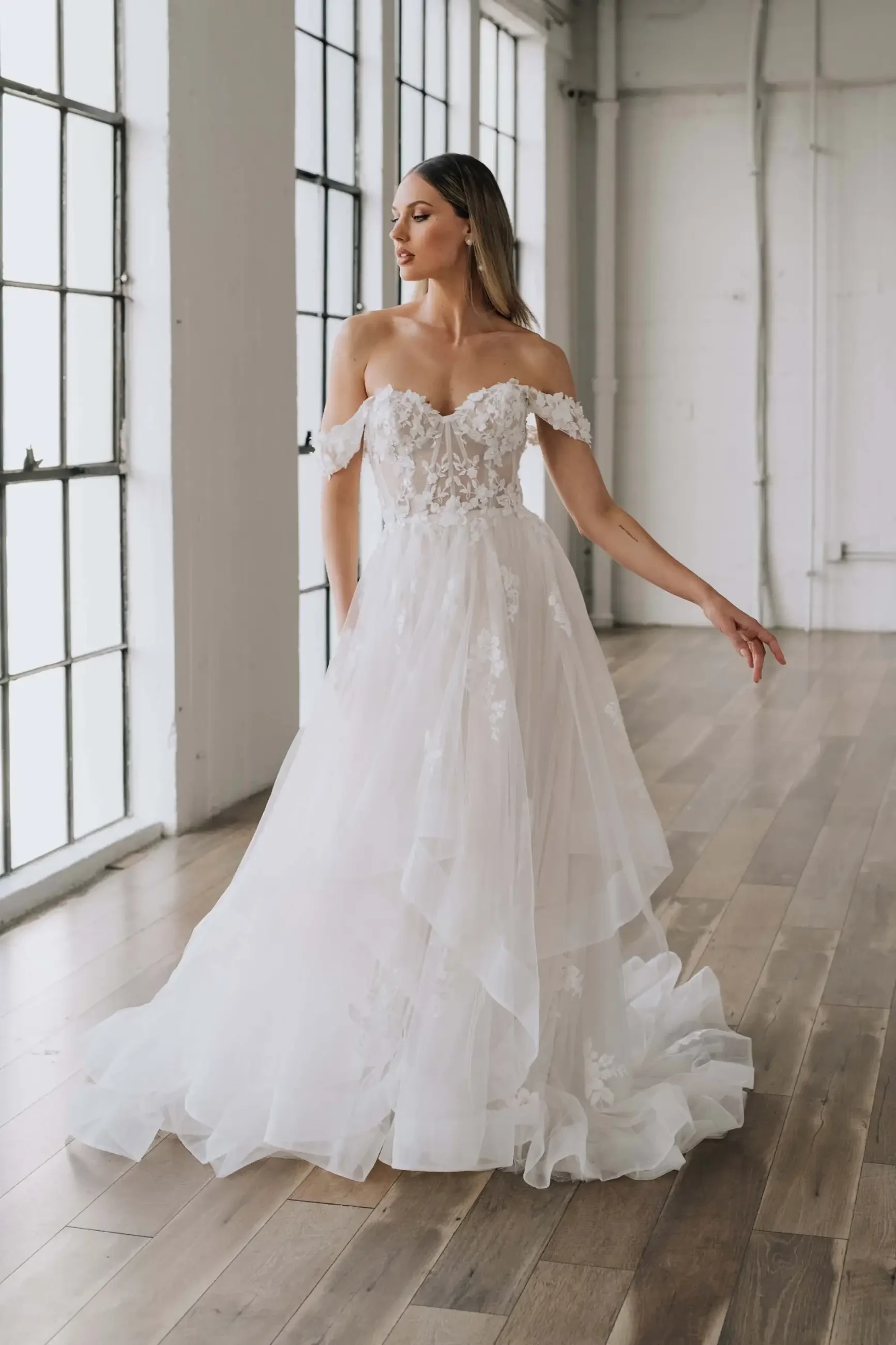 A woman in a strapless, off-the-shoulder wedding dress with floral embellishments, posing gracefully indoors near large windows.