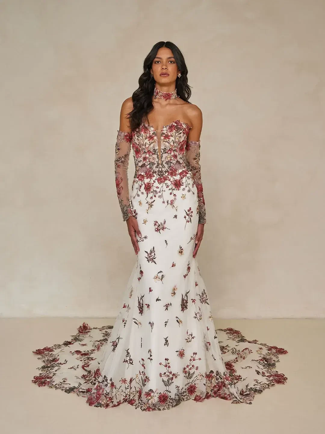 A woman is wearing an elegant white gown adorned with floral embroidery, featuring off-the-shoulder sleeves and a train. The dress has intricate detailing with shades of red and green.