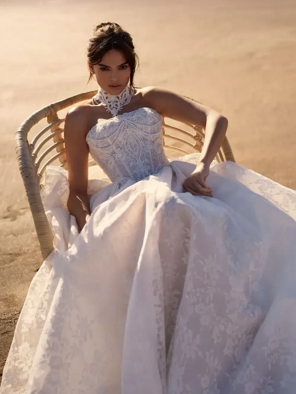 A woman in a strapless wedding dress with intricate lace details sitting on a stylish chair in a sandy environment.