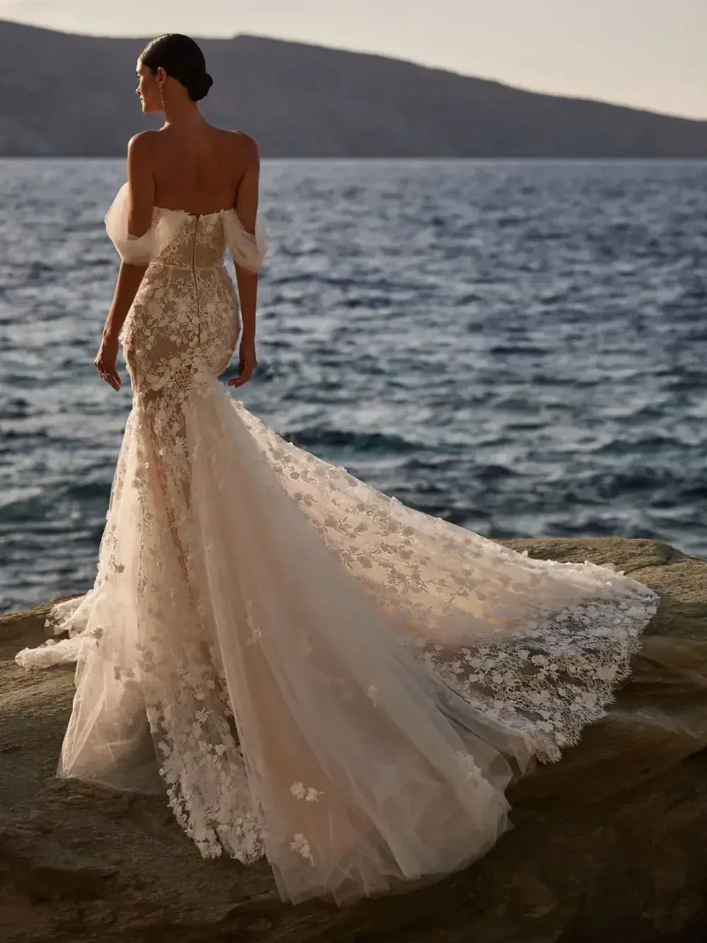A woman wearing an elegant wedding dress with floral details stands near the water, facing away.