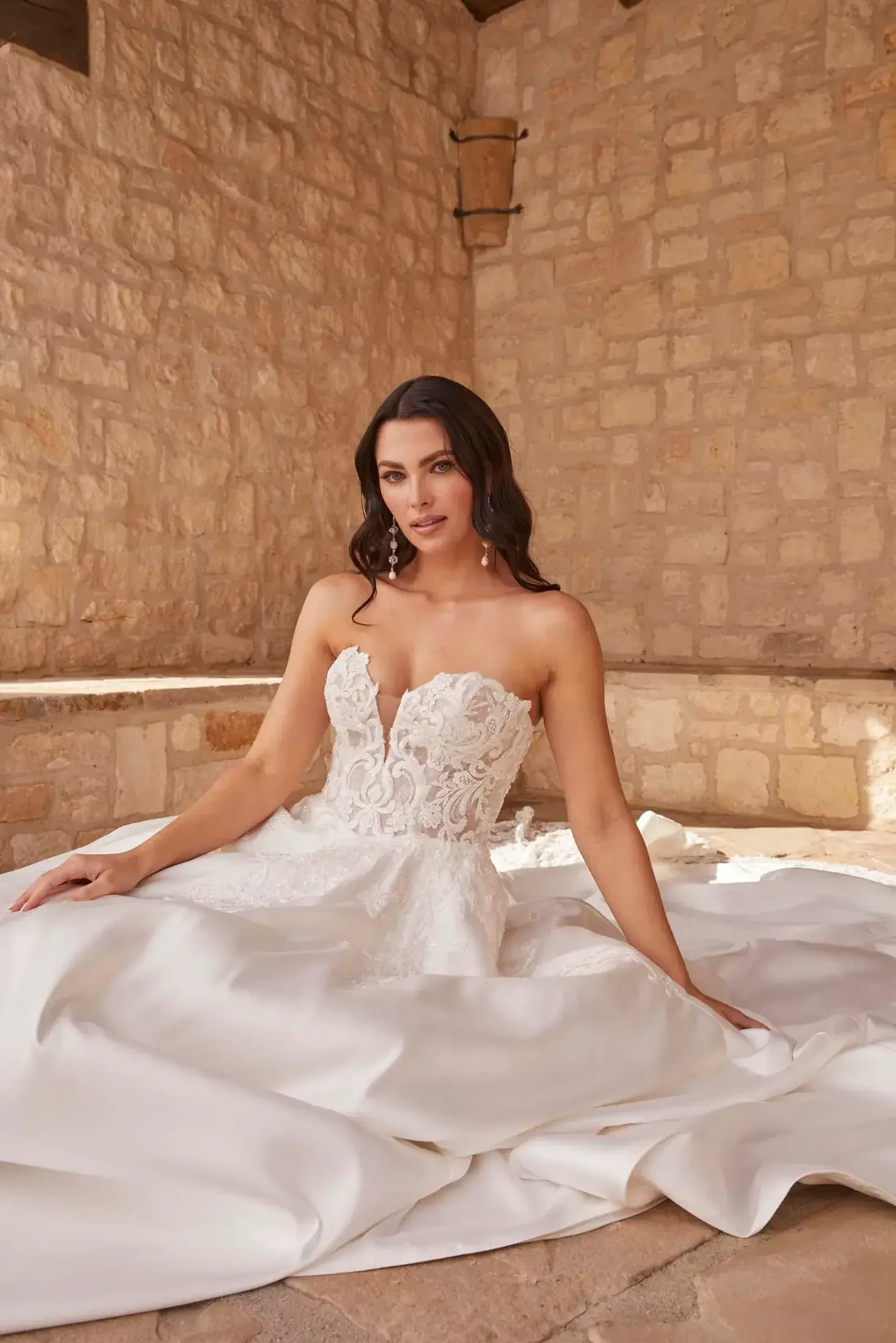 A woman in a strapless white wedding dress is seated on a stone surface with her legs gracefully arranged. The dress features intricate lace detailing at the bodice and flows elegantly around her. She has long, dark hair styled in soft waves and is looking directly at the camera, exuding confidence and poise. The background consists of warm stone walls that complement her bridal look.