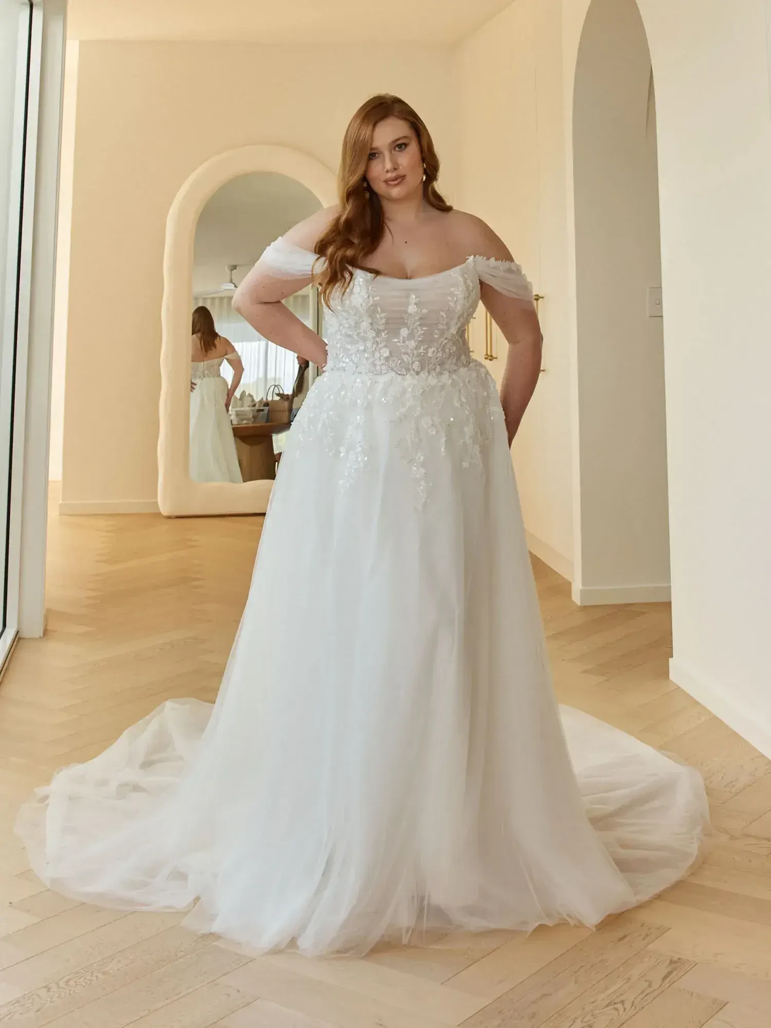 A plus-size woman wearing an off-the-shoulder wedding dress, standing in a bright room with a mirror and soft lighting. 
